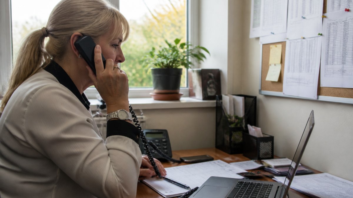 A school business manager on the phone in a small office, rota sheets pinned to a board and a laptop showing a staff absence list on the desk, morning light through the window