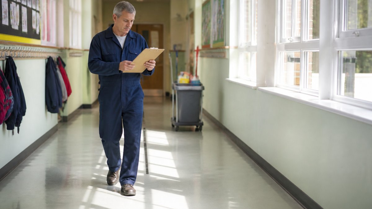 A school caretaker in blue overalls checking a clipboard while walking through a school corridor, cleaning trolley visible in the background, morning light through windows